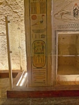 Ancient hieroglyphics on a weathered stone wall, featuring symbols such as birds and a sun disk. The stone surface is textured and appears aged, with carved and painted imagery. A glass barrier is in place, suggesting preservation or display.