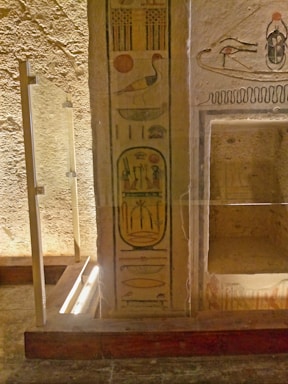 Ancient hieroglyphics on a weathered stone wall, featuring symbols such as birds and a sun disk. The stone surface is textured and appears aged, with carved and painted imagery. A glass barrier is in place, suggesting preservation or display.