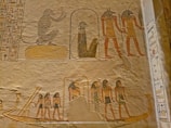 Ancient Egyptian wall reliefs featuring depictions of various figures, including humanoid deities with animal heads. A seated figure is shown alongside others in a standing posture, likely signifying a narrative or ceremonial scene. The carvings exhibit detailed hieroglyphics surrounding the images.