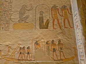 Ancient Egyptian wall reliefs featuring depictions of various figures, including humanoid deities with animal heads. A seated figure is shown alongside others in a standing posture, likely signifying a narrative or ceremonial scene. The carvings exhibit detailed hieroglyphics surrounding the images.