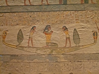 An ancient Egyptian wall carving depicting three human figures on a boat. The central figure resembles a deity or a significant personage, with distinct headwear, flanked by two figures on either side. The boat is adorned with depictions of serpents at both ends, and there are hieroglyphics above and below the scene, suggesting a religious or ceremonial significance.