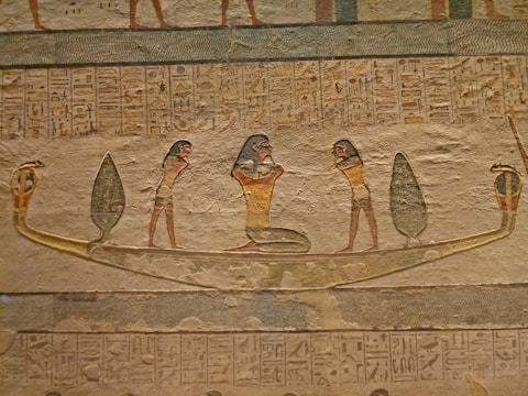 An ancient Egyptian wall carving depicting three human figures on a boat. The central figure resembles a deity or a significant personage, with distinct headwear, flanked by two figures on either side. The boat is adorned with depictions of serpents at both ends, and there are hieroglyphics above and below the scene, suggesting a religious or ceremonial significance.