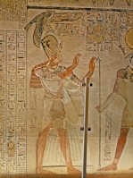 An ancient Egyptian wall painting featuring a figure in traditional attire, with a headdress and a long skirt, raising their hands in what appears to be a gesture of worship or communication. Hieroglyphics are inscribed on the wall surrounding the figure. The artwork is rich in detail and coloration, indicative of Egyptian artistic style.