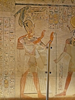 An ancient Egyptian wall painting featuring a figure in traditional attire, with a headdress and a long skirt, raising their hands in what appears to be a gesture of worship or communication. Hieroglyphics are inscribed on the wall surrounding the figure. The artwork is rich in detail and coloration, indicative of Egyptian artistic style.