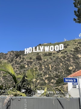 The iconic Hollywood sign is prominently displayed on a hillside with a clear blue sky in the background. In the foreground, palm leaves and a street sign labeled Deronda Dr are visible, adding a suburban touch to the scene.