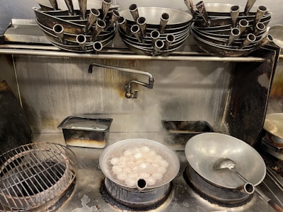 A kitchen scene with a metal stove and various cooking utensils. There is a pot of boiling water containing eggs and steam rising from it. On the stove, there are several stacks of metal ladles and a faucet above the boiling pot. A metal rack, a soapy tray, and a large ladle resting in a bowl are also visible.