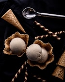 Two scoops of ice cream sit in waffle bowls surrounded by waffle cones and chocolate-filled wafer rolls on a dark surface. An ice cream scoop lies nearby, contributing to the arrangement.