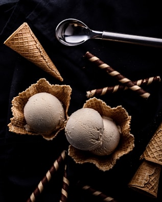 Two scoops of ice cream sit in waffle bowls surrounded by waffle cones and chocolate-filled wafer rolls on a dark surface. An ice cream scoop lies nearby, contributing to the arrangement.
