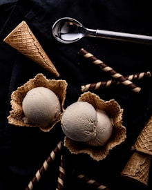 Two scoops of ice cream sit in waffle bowls surrounded by waffle cones and chocolate-filled wafer rolls on a dark surface. An ice cream scoop lies nearby, contributing to the arrangement.