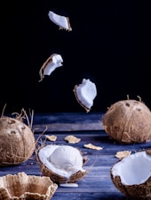 An artistic display of coconuts and ice cream on a dark wooden surface. Two whole coconuts and several halves are arranged alongside a scoop of vanilla ice cream placed in a coconut shell. Fragments of wafers are scattered around, with a few coconut pieces suspended in the air, adding a dynamic element.