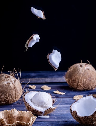 An artistic display of coconuts and ice cream on a dark wooden surface. Two whole coconuts and several halves are arranged alongside a scoop of vanilla ice cream placed in a coconut shell. Fragments of wafers are scattered around, with a few coconut pieces suspended in the air, adding a dynamic element.