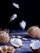 An artistic display of coconuts and ice cream on a dark wooden surface. Two whole coconuts and several halves are arranged alongside a scoop of vanilla ice cream placed in a coconut shell. Fragments of wafers are scattered around, with a few coconut pieces suspended in the air, adding a dynamic element.
