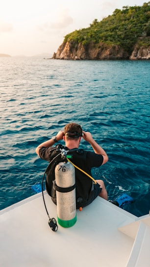 A person is seated on the edge of a boat, wearing scuba diving gear and preparing to enter the water. The sea spreads out in front, with the blue water contrasting against a rocky, green-covered island in the distance.