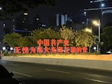 Bright red Chinese characters are prominently displayed on a street-side structure, surrounded by lush green trees. The setting is urban, with buildings and glowing streetlights visible in the background during nighttime.