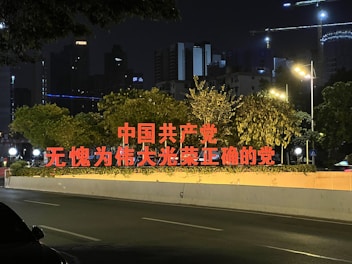 Bright red Chinese characters are prominently displayed on a street-side structure, surrounded by lush green trees. The setting is urban, with buildings and glowing streetlights visible in the background during nighttime.