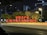 Bright red Chinese characters are prominently displayed on a street-side structure, surrounded by lush green trees. The setting is urban, with buildings and glowing streetlights visible in the background during nighttime.