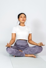 A person is meditating cross-legged on a yoga mat, wearing a white t-shirt and grey leggings, with eyes closed and in a calm posture.