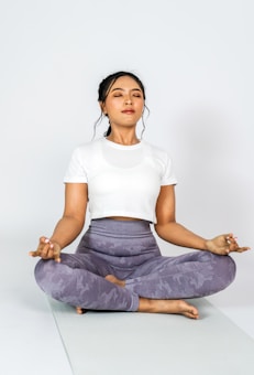 A person is meditating cross-legged on a yoga mat, wearing a white t-shirt and grey leggings, with eyes closed and in a calm posture.