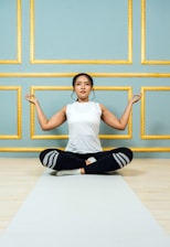 A person is seated cross-legged on a yoga mat, performing a meditation pose with closed eyes. The background features an elegant wall with teal panels and gold borders.