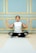 A person is seated cross-legged on a yoga mat, performing a meditation pose with closed eyes. The background features an elegant wall with teal panels and gold borders.