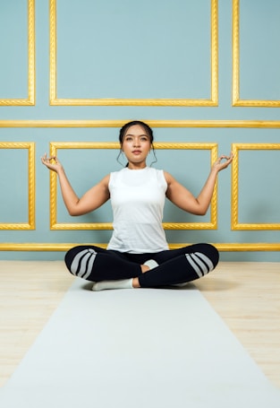 A person is seated cross-legged on a yoga mat, performing a meditation pose with closed eyes. The background features an elegant wall with teal panels and gold borders.