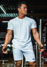 A muscular man is standing in a gym setting, wearing a white T-shirt and white shorts. His arms are prominently showing veins and he is looking to the side, appearing focused and intense. The gym environment is visible in the background with gym equipment and weights.