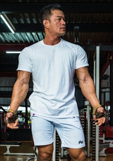 A muscular man is standing in a gym setting, wearing a white T-shirt and white shorts. His arms are prominently showing veins and he is looking to the side, appearing focused and intense. The gym environment is visible in the background with gym equipment and weights.