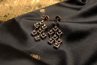 Elegant geometric earrings made of a golden material embellished with small sparkling stones are placed on a textured dark fabric. The background features a subtle gold pattern, adding a touch of luxury to the composition.