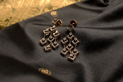 Elegant geometric earrings made of a golden material embellished with small sparkling stones are placed on a textured dark fabric. The background features a subtle gold pattern, adding a touch of luxury to the composition.