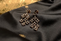Elegant geometric earrings made of a golden material embellished with small sparkling stones are placed on a textured dark fabric. The background features a subtle gold pattern, adding a touch of luxury to the composition.