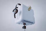 A white cube-like object with perforated patterns is mounted on a stand. It has a leather strap attached to the top and displays the text 'easybadwork.' A set of black headphones with a cartoon character sticker featuring blue hair and sunglasses is hanging from the cube.