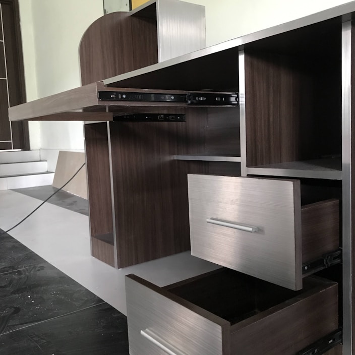 A partially assembled wooden cabinet with several open drawers and compartments, designed with a modern aesthetic. The cabinet features a sleek, dark wood finish combined with metallic elements, and is situated in a room with steps leading to another area.