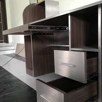 A partially assembled wooden cabinet with several open drawers and compartments, designed with a modern aesthetic. The cabinet features a sleek, dark wood finish combined with metallic elements, and is situated in a room with steps leading to another area.