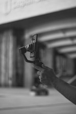 A hand holding a smartphone mounted on a stabilizing gimbal in an urban environment. The image is in black and white, focusing on the technological gadget.