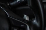 A close-up of a car's interior focusing on a metallic paddle shifter attached to the steering wheel. The shifter has a clear plus sign engraved on it, and the surrounding areas appear to be composed of dark leather and plastic elements.