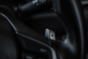 A close-up of a car's interior focusing on a metallic paddle shifter attached to the steering wheel. The shifter has a clear plus sign engraved on it, and the surrounding areas appear to be composed of dark leather and plastic elements.