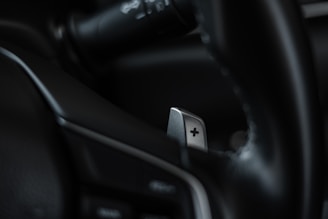 A close-up of a car's interior focusing on a metallic paddle shifter attached to the steering wheel. The shifter has a clear plus sign engraved on it, and the surrounding areas appear to be composed of dark leather and plastic elements.