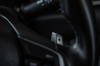 A close-up of a car's interior focusing on a metallic paddle shifter attached to the steering wheel. The shifter has a clear plus sign engraved on it, and the surrounding areas appear to be composed of dark leather and plastic elements.