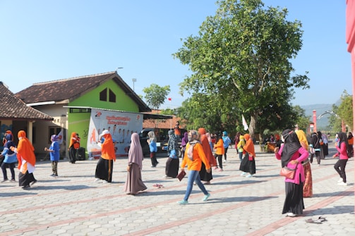 A group of people, mostly women, are engaging in a physical activity outdoors. They are dressed in colorful attire, with many wearing orange or pink. The setting is in an open area with a paved surface near buildings with traditional roof designs. A large tree provides some shade. A banner with text and images is visible in the background.