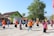 A group of people, mostly women, are engaging in a physical activity outdoors. They are dressed in colorful attire, with many wearing orange or pink. The setting is in an open area with a paved surface near buildings with traditional roof designs. A large tree provides some shade. A banner with text and images is visible in the background.