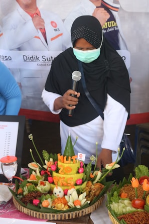 A person wearing a black headscarf and a mask is holding a microphone while standing next to a large traditional Indonesian food arrangement featuring colorful vegetables and rice shaped into a cone. The food display includes carvings that read 'HUT 20 PKS' and is surrounded by flowers and other decorative elements.