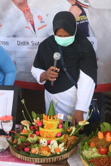 A person wearing a black headscarf and a mask is holding a microphone while standing next to a large traditional Indonesian food arrangement featuring colorful vegetables and rice shaped into a cone. The food display includes carvings that read 'HUT 20 PKS' and is surrounded by flowers and other decorative elements.