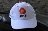 A white baseball cap is positioned on a dark textured surface. The cap features an orange circular logo with a crescent shape and the letters 'PKS' written beneath it in black. The background is blurred and dark, adding contrast to the cap.