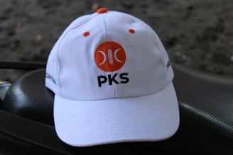 A white baseball cap is positioned on a dark textured surface. The cap features an orange circular logo with a crescent shape and the letters 'PKS' written beneath it in black. The background is blurred and dark, adding contrast to the cap.