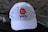 A white baseball cap is positioned on a dark textured surface. The cap features an orange circular logo with a crescent shape and the letters 'PKS' written beneath it in black. The background is blurred and dark, adding contrast to the cap.