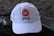 A white baseball cap is positioned on a dark textured surface. The cap features an orange circular logo with a crescent shape and the letters 'PKS' written beneath it in black. The background is blurred and dark, adding contrast to the cap.