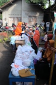 A group of people are gathered outdoors near a table filled with plastic bags, papers, and a bottle of water. Several individuals are wearing orange clothing and headscarves. In the background, there is a row of parked motorcycles and a building with a textured wall and wooden doors.