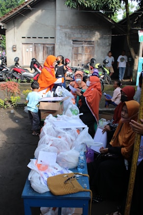 A group of people are gathered outdoors near a table filled with plastic bags, papers, and a bottle of water. Several individuals are wearing orange clothing and headscarves. In the background, there is a row of parked motorcycles and a building with a textured wall and wooden doors.