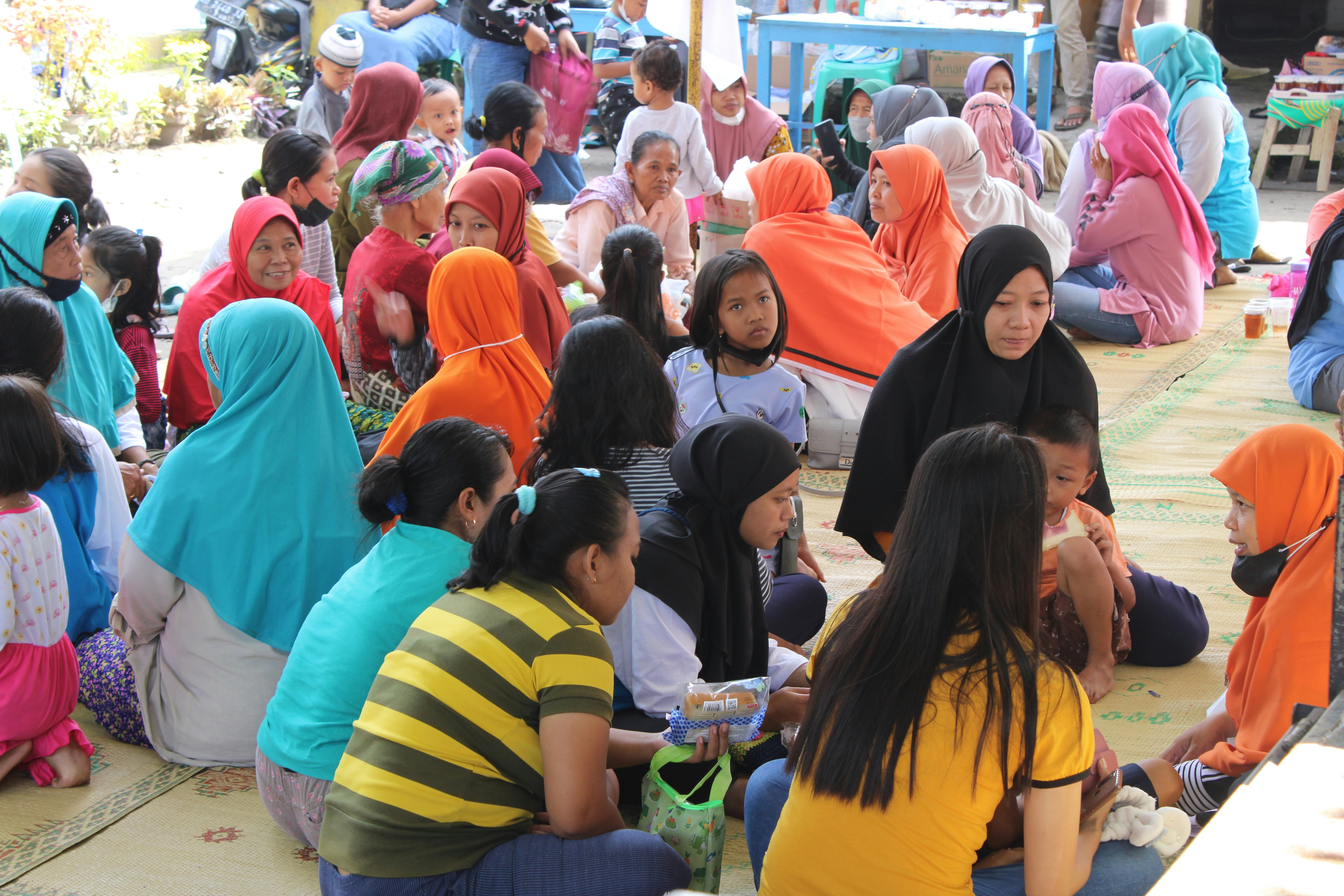 A large group of people gathered together, predominantly consisting of women and children, many of whom are wearing colorful hijabs and clothing. They are seated on mats in an outdoor setting, possibly engaged in conversation or waiting for an event or activity.