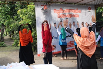A group of people wearing colorful headscarves and shirts are standing outdoors with their arms raised, seemingly participating in an exercise or stretching activity. They are in front of a large banner featuring images and text. The setting includes greenery and a covered area.